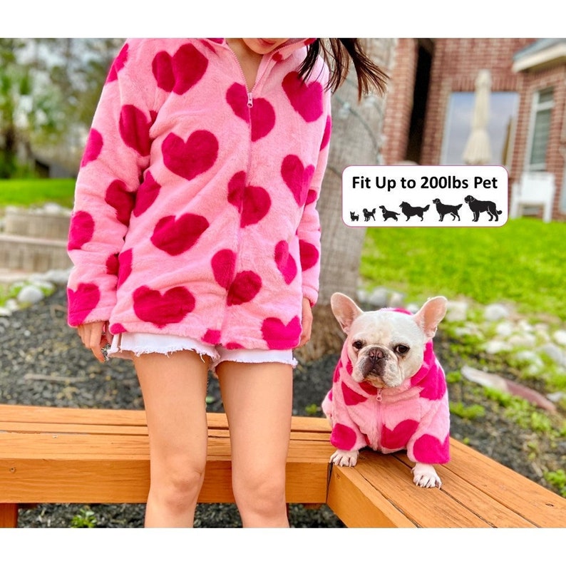 Puede incluir: Una chaqueta de forro polar rosa con estampado de corazones y cierre de cremallera. Un perro peque&ntilde;o que lleva una chaqueta de forro polar rosa a juego con estampado de corazones est&aacute; sentado en un banco de madera. El texto "Fit Up to 200lbs Pet" es visible en la imagen.