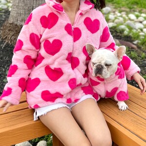 Puede incluir: Una chaqueta de forro polar rosa con estampado de corazones y una chaqueta de forro polar rosa a juego para un perro. La chaqueta tiene una cremallera y una capucha. El perro es un Bulldog Franc&eacute;s.