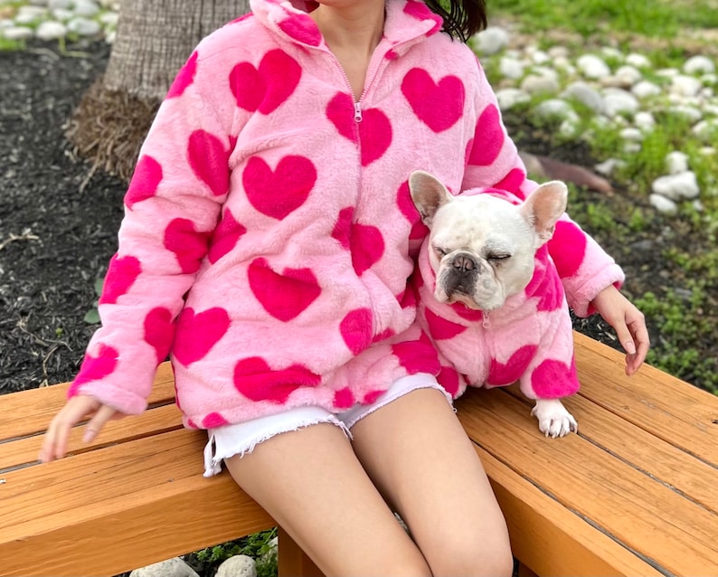 Puede incluir: Una chaqueta de forro polar rosa con estampado de corazones y una chaqueta de forro polar rosa a juego para un perro. La chaqueta tiene una cremallera y una capucha. El perro es un Bulldog Franc&eacute;s.