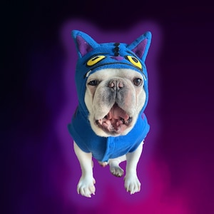 May include: A white French Bulldog wearing a blue cat costume with purple ears and yellow eyes. The dog is centered, facing forward, with its mouth open. The background is a gradient of purple and black.