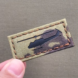 Peut inclure: Un écusson rectangulaire avec un motif de camouflage dans les tons de vert, marron et beige. L'écusson présente une silhouette noire d'un hélicoptère. Les bords sont soulignés par des coutures marron.