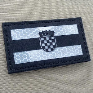 Può includere: Un patch della bandiera croata in bianco e nero con una superficie riflettente. Il patch è cucito su uno sfondo in tessuto beige.
