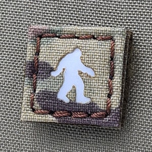 May include: A small, square patch with a brown, tan, and white camouflage pattern. The patch has a white outline and a white silhouette of a Bigfoot inside.