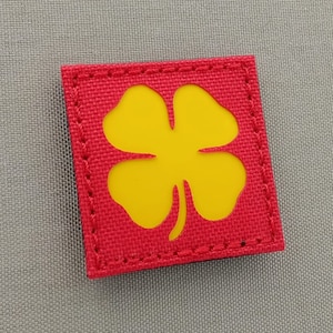 Può includere: Una toppa quadrata rossa con un design a quadrifoglio giallo. La toppa ha un bordo cucito.