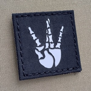 May include: Black fabric patch with a white skeleton hand making the 'W' sign. The patch has a stitched border.