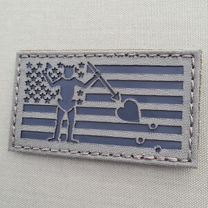 Könnte beinhalten: Ein gestickter Patch in Schwarz und Grau mit einer Silhouette einer Person, die eine Waffe und ein Herz hält, überlagert mit einem amerikanischen Flaggenmuster.