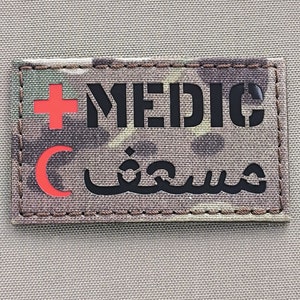 Könnte beinhalten: Ein Patch aus Tarnstoff mit dem Wort "MEDIC" in schwarzen Buchstaben, einem roten Kreuz, einem roten Halbmond und dem arabischen Wort für "Sanitäter" in schwarzen Buchstaben.