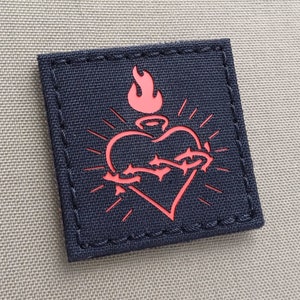 May include: A dark blue patch with a red embroidered heart with a crown and thorns. The heart is surrounded by a red flame.
