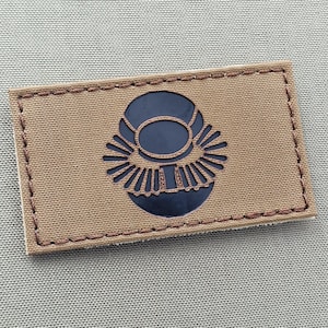 May include: A brown fabric patch with a black and white stylized design of a circle with a smaller circle inside and radiating lines around the outer circle.