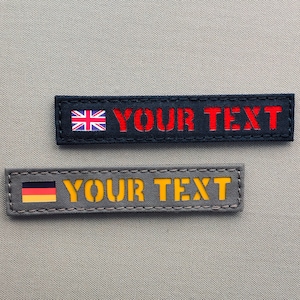 May include: Two black and gray fabric patches with white stitching. The top patch has a red, white, and blue flag and the text "YOUR TEXT" in red. The bottom patch has a black, yellow, and red flag and the text "YOUR TEXT" in yellow.