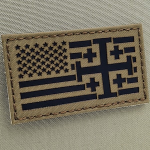 Könnte beinhalten: Ein brauner Patch mit einer schwarzen gestickten amerikanischen Flagge und einem schwarzen Kreuzmuster. Der Patch hat einen gestickten Rand.