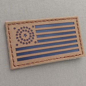 Könnte beinhalten: Ein brauner Stoff-Patch mit einem schwarzen amerikanischen Flaggen-Design. Die Flagge hat einen Kreis aus Sternen im Kanton und 13 Streifen.
