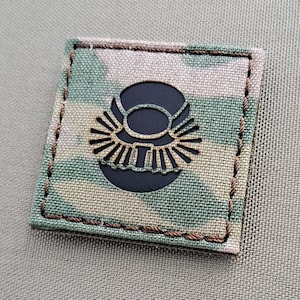 May include: A black and brown camouflage fabric patch with a black embroidered symbol of a circle with a wing design inside.