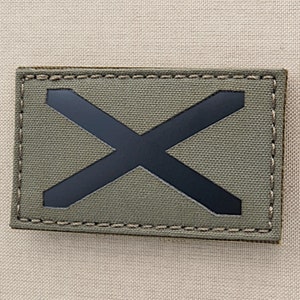 Könnte beinhalten: Ein grüner Stoff-Patch mit einem schwarzen X, das darauf genäht ist. Der Patch hat einen genähten Rand.