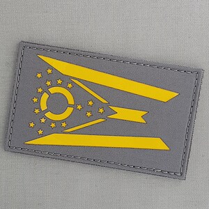 Può includere: Una toppa grigia con un disegno ricamato giallo della bandiera della città di Cincinnati, Ohio. La bandiera presenta un cerchio bianco con una stella a tre punte all'interno, circondata da un anello di 13 stelle a cinque punte.