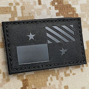 Pode incluir: Um patch retangular preto com um design de bandeira estilizado. A bandeira inclui duas estrelas e uma forma retangular, com listras diagonais. O patch está sobre um fundo de camuflagem.