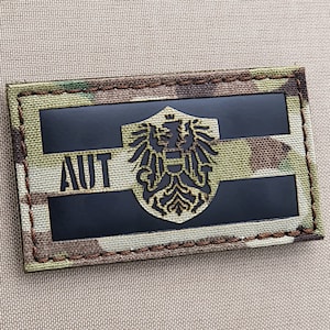 May include: A black and white embroidered patch featuring the Austrian flag and the text "AUT". The patch is set against a camouflage background.