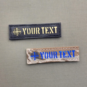Op de afbeelding: Twee stoffen patches in zwart en bruin met een kruis en de tekst "YOUR TEXT" in gouden en blauwe letters.