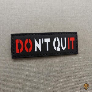 Op de afbeelding: Een rechthoekige zwarte patch met gestikte randen met de tekst DON'T QUIT in een vetgedrukt sjabloonlettertype. De letters D, O en N zijn rood, terwijl de letters T, Q, U, I en T wit zijn, wat zorgt voor een ontwerp met een hoog contrast.