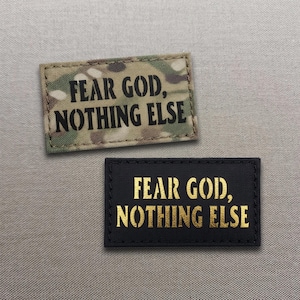 以下が含まれることがあります： 「FEAR GOD, NOTHING ELSE」という文字が記された2つの長方形のパッチ。1つは黒い文字の迷彩柄で、もう1つはメタリックゴールドの文字が施された無地の黒色です。どちらのパッチも縁が丁寧に縫製されています。