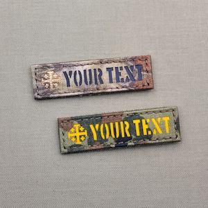 Puede incluir: Dos parches rectangulares de tela con patrón de camuflaje y bordes cosidos. El parche superior presenta un icono de cruz color cobre y el texto "YOUR TEXT" en azul oscuro. El parche inferior muestra un icono de cruz amarilla y "YOUR TEXT" en amarillo.