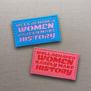 May include: Two rectangular patches with the text "WELL-BEHAVED WOMEN RARELY MAKE HISTORY". One patch is blue with pink text, the other is pink with red text. Both have stitched edges.