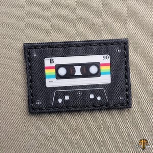 May include: Black rectangular patch featuring a retro cassette tape design. The tape has a white label with the text "SIDE B" and "90", and a rainbow stripe. The patch is stitched with black thread.