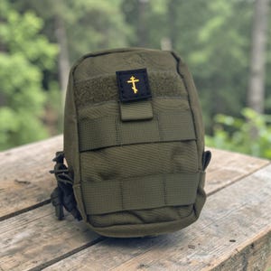 K&ouml;nnte beinhalten: Olivgr&uuml;ne taktische Tasche mit einem schwarzen und goldenen Kreuzaufn&auml;her. Die Tasche hat mehrere Riemen und einen Klettverschluss. Der Hintergrund ist eine verschwommene Ansicht einer Holzoberfl&auml;che und gr&uuml;ner Bl&auml;tter.