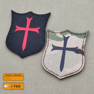 May include: Two embroidered patches in the shape of a shield with a cross inside. The patch on the left is black with a red cross. The patch on the right is a camouflage pattern with a black cross. The text on the bottom of the image reads "MODEL SHOWN", "SIZE 3"x2.25", "CHOOSE FROM +700 COLOR OPTIONS".