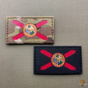 May include: Two rectangular patches depicting the Florida state flag. One has a camouflage background with red detailing, the other is black with red detailing. Both patches feature the state seal at the centre.