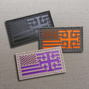 Può includere: Tre toppe rettangolari con un design a bandiera stilizzato, caratterizzato da un campo di stelle e strisce orizzontali integrate con un simbolo a croce. Una toppa è nera, una arancione brillante e una viola, tutte con bordi cuciti.