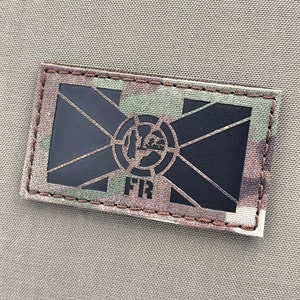 May include: Black and white patch with a stylized design of a cross and a circle with a symbol inside. The text "FIR" is below the design. The patch is sewn onto a camouflage fabric.