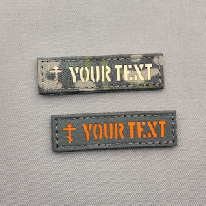 Op de afbeelding: Twee rechthoekige patches met de tekst "YOUR TEXT". Eén patch heeft een camouflagepatroon met gouden tekst en een kruissymbool. De andere patch is grijs met oranje tekst en een kruissymbool.