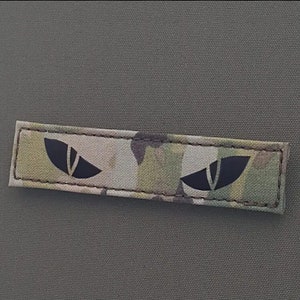 Op de afbeelding: Een camouflage-stofpatch met twee zwarte oogvormige ontwerpen. De patch heeft een gestikte rand.