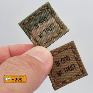 May include: Two square patches with the phrase "In God We Trust" embroidered in black thread. The patches are made of different camouflage fabrics. One patch is green and brown, the other is brown.