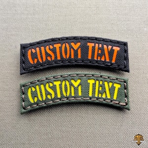 Può includere: Due toppe con testo personalizzato. Una è nera con testo arancione, l'altra è verde con testo giallo. Entrambe le toppe hanno il testo "CUSTOM TEXT" in un carattere grassetto, simile a uno stencil. Le toppe sono arcuate e hanno bordi cuciti.