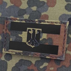 Puede incluir: Un parche rectangular con la bandera alemana y el emblema del águila, cosido sobre tela de camuflaje. La tela presenta un patrón de marrón, verde y negro. El diseño de la bandera incluye rayas horizontales negras.