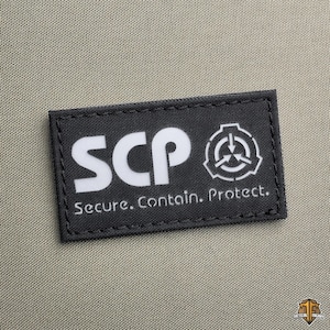 Può includere: Una toppa rettangolare nera con testo bianco e un simbolo. Il testo recita "SCP Secure. Contain. Protect." Il simbolo è un cerchio bianco con tre frecce rivolte verso l'interno. La toppa è su un tessuto beige.