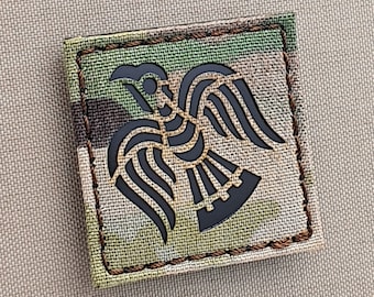 May include: A black and white embroidered patch with a stylized raven design on a camouflage background. The patch has a stitched border.