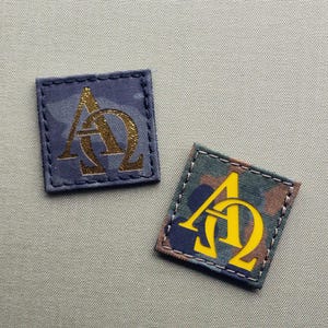 May include: Two square patches with a camouflage pattern. The patches have a gold "AΩ" symbol on them. One patch is blue and the other is green.