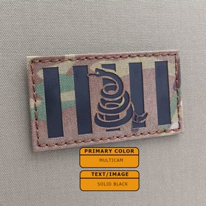 May include: A black and green camouflage patch with a black snake with a coiled tail and a 'Don't Tread On Me' design. The patch is sewn onto a tan fabric background.