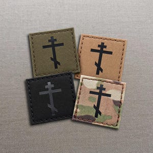 Könnte beinhalten: Vier quadratische Aufnäher mit genähten Rändern, die jeweils ein schwarzes orthodoxes Kreuz zeigen. Die Aufnäher sind in Olivgrün, Beige, Schwarz und einem mehrfarbigen Tarnmuster erhältlich und bestehen aus einem strapazierfähigen, strukturierten Stoff.