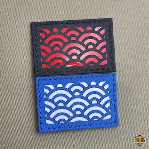 Könnte beinhalten: Zwei rechteckige Aufnäher mit einem Wellenmuster. Der obere Aufnäher ist schwarz mit einem roten Wellendesign, und der untere Aufnäher ist blau mit einem weißen Wellendesign. Beide Aufnäher sind mit einem Kontrastfaden genäht.