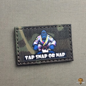 Può includere: Toppa rettangolare con motivo mimetico e un gorilla dei cartoni animati con una divisa di arti marziali. Il testo "TAP SNAP OR NAP" è in basso. La toppa è bordata con cuciture nere.