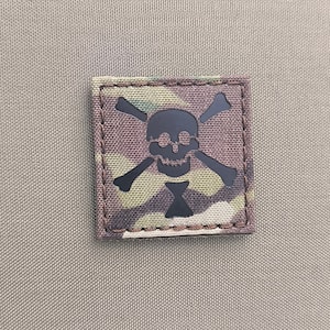 May include: A black skull and crossbones patch with a camouflage background. The patch is sewn onto a beige fabric.