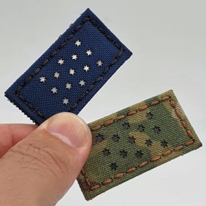 Può includere: Sono mostrate due toppe rettangolari, una blu navy e una mimetica. Ogni toppa presenta ritagli a forma di stella ed è bordata da cuciture a contrasto. La toppa blu navy ha cuciture nere, mentre la toppa mimetica ha cuciture marroni.