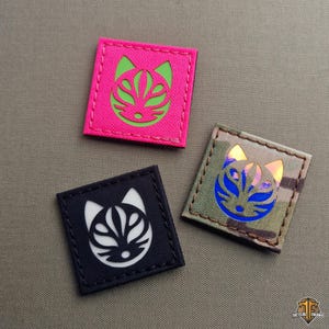 May include: Three square patches with a cat face design. One patch is hot pink with a green cat face, another is black with a white cat face, and the third is camouflage with a blue cat face.