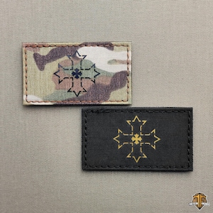 Op de afbeelding: Twee rechthoekige patches. Eén met een camouflagepatroon met een zwart en blauw kruisontwerp. De andere is zwart met een gouden kruisontwerp. Beide hebben gestikte randen, waarschijnlijk voor tactische uitrusting.