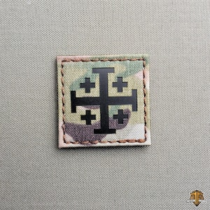 Op de afbeelding: Een vierkante patch met een zwart Jeruzalemkruisontwerp op een camouflage achtergrond. De patch is omrand met bruine stiksels en geplaatst tegen een neutrale, getextureerde achtergrond.