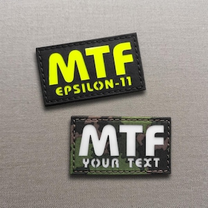 Pode incluir: Duas etiquetas retangulares. A etiqueta superior é preta com as letras "MTF" em amarelo brilhante e o texto "EPSILON-11". A etiqueta inferior tem um padrão de camuflagem com as letras "MTF" em branco e o texto "YOUR TEXT".